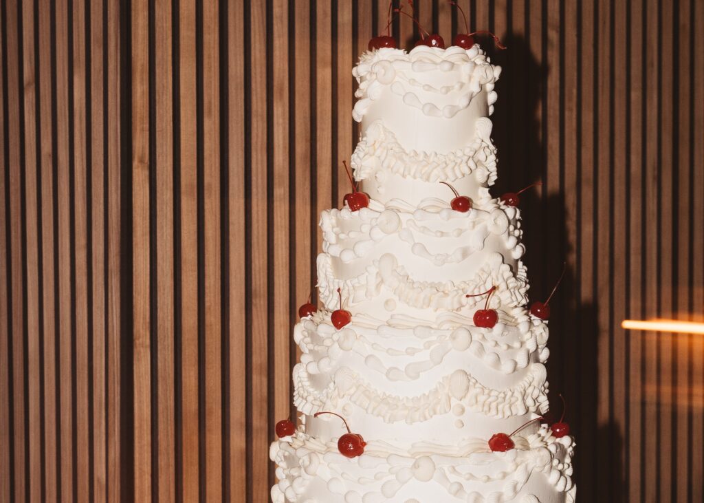 A vintage style wedding cake covered in white frosting and cherries. 
