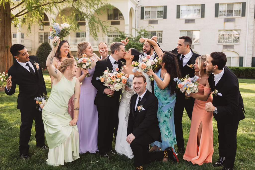 Bride and groom kiss with their wedding party, dressed in spring pastels cheering them on, on the front lawn of the Madison Hotel in New Jersey. Taken by NJ wedding photographer Madison Neumann Photography.