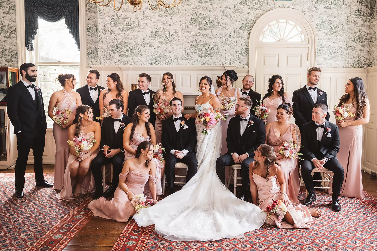Bride and groom with their wedding party inside venue with decorative wallpaper and rugs. Bridesmaids wear light pink gowns and groomsmen wear black tuxedos. Photographed by South Jersey, Philadelphia based wedding photographer Madison Neumann Photography.
