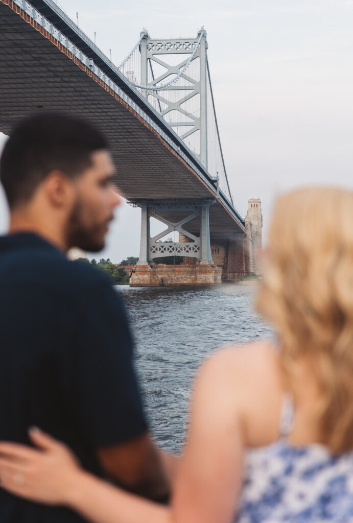 A couple looks at one another with the Ben Franklin bridge in the background. They are standing on Race Street Pier in Philadelphia. Taken by Philadelphia wedding photographer Madison Neumann Photography. 
