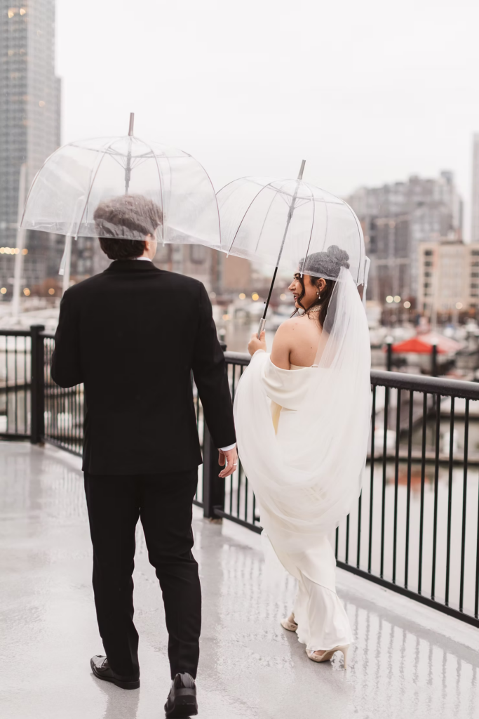 Bride and groom walk together outside in rain holding clear umbrellas. Wedding day at Maritime Parc in Jersey City, NJ. Photographed by South Jersey, Philadelphia based wedding photographer Madison Neumann Photography.