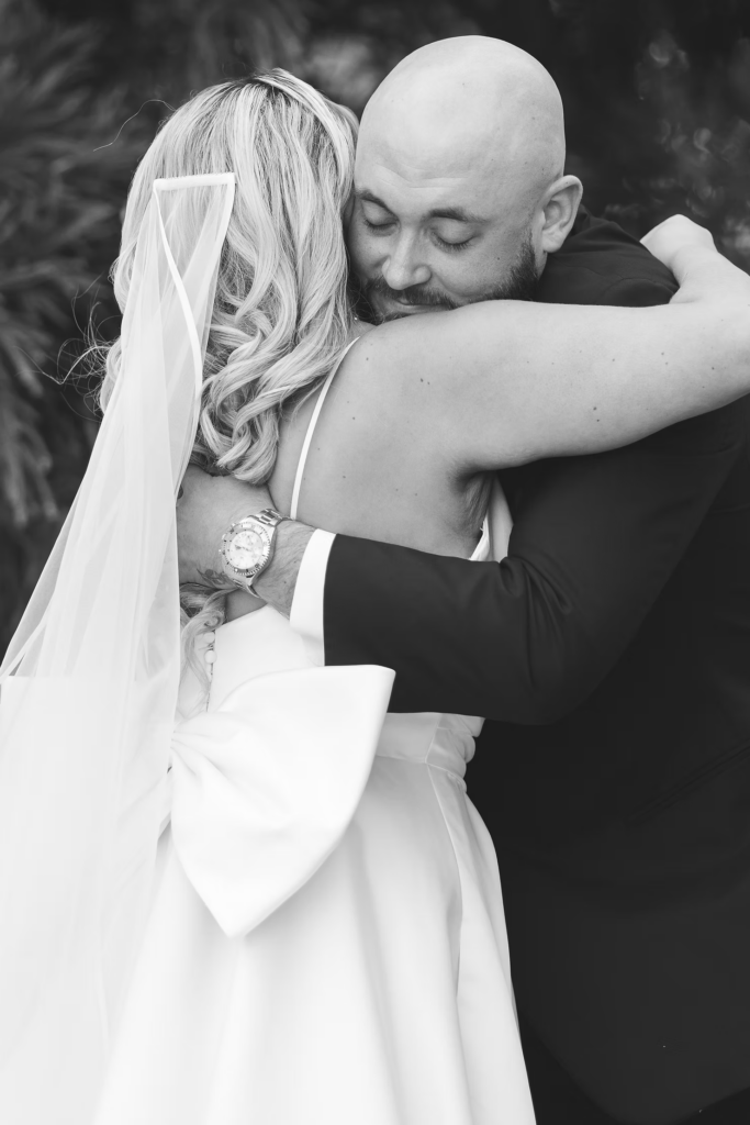 Black and white photograph of bride and groom embracing after first look moment on wedding day at The Ivy at Ellis Preserve in Newtown Square, PA. Bride in gown with bow on back and long veil, groom in tux wearing watch. Portrait photographed by South Jersey, Pennsylvania region wedding photographer Madison Neumann Photography.