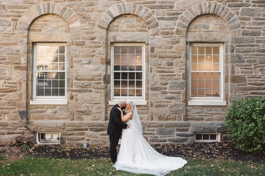 Bride and groom outside stone building with windows pose for wedding portrait at for wedding day at The Ivy at Ellis Preserve in Newtown Square, PA by Finley Catering. Photographed by South Jersey, Pennsylvania region wedding photographer Madison Neumann Photography.