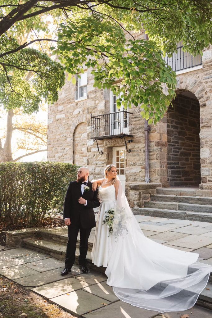 Wedding portrait of bride and groom posing outside stone building framed by tree. Bride in gown with bouquet and long veil, groom in tuxedo. Day photographed by South Jersey, Pennsylvania region wedding photographer Madison Neumann Photography at The Ivy at Ellis Preserve in Newtown Square, PA.