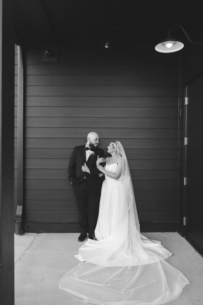 Bride with bouquet and long veil standing with groom outside modern barn venue for wedding day in Newtown Square, PA. Wedding day at The Ivy at Ellis Preserve photographed by South Jersey, Pennsylvania region wedding photographer Madison Neumann Photography.