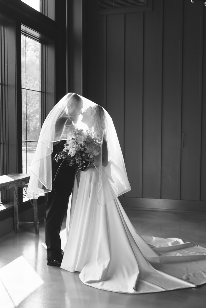 Black and white sunlit portrait of bride and groom with long flowing veil and large bouquet, bride in gown with train. Photographed for wedding day at The Ivy at Ellis Preserve in Newtown Square, PA by South Jersey, Pennsylvania region wedding photographer Madison Neumann Photography.
