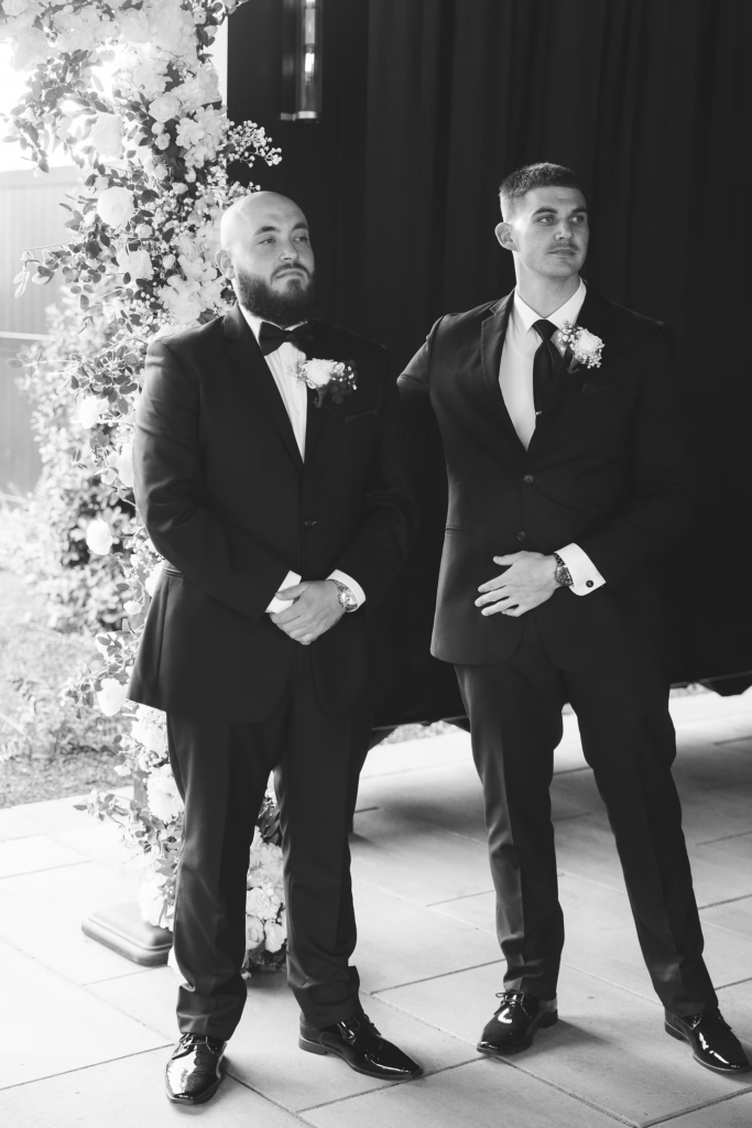 Sunlit black and white portrait of groom watching bride walk down the aisle during wedding ceremony at The Ivy at Ellis Preserve by Finley Catering in Newtown Square, PA. Photographed by South Jersey, Pennsylvania region wedding photographer Madison Neumann Photography.