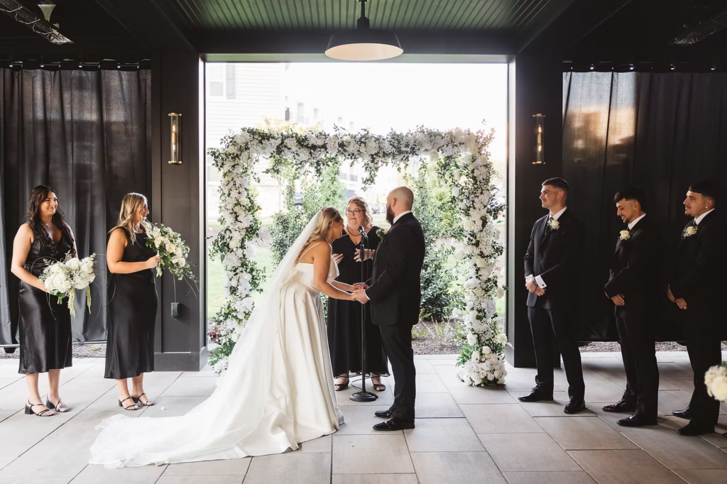 Bride and groom hold hands in front of floral arbor, sharing a laugh during exchanging of vows during wedding ceremony at The Ivy at Ellis Preserve by Finley Catering in Newtown Square, PA. Photographed by South Jersey, Pennsylvania region wedding photographer Madison Neumann Photography.