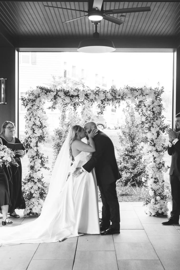 Bride and groom seal wedding vows in front of flower arbor at the altar for wedding ceremony at The Ivy at Ellis Preserve by Finley Catering in Newtown Square, PA. Photographed by South Jersey, Pennsylvania region wedding photographer Madison Neumann Photography.