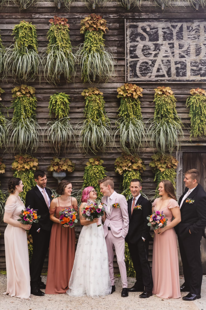 Wedding party photo at Terrain in Glen Mills, PA. Taken in front of a plant wall on the property, wooden and covered in green plants. The wedding party wears mismatched shades of pink. Taken by philly wedding photographer Madison Neumann Photography.