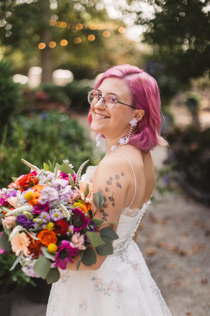 Bridal portrait at Terrain in Glen Mills, PA taken in the garden. She wears a white dress with pink flowers and her hair is a matching shade of pink. She holds a colorful bouquet. Taken by philly wedding photographer Madison Neumann Photography.