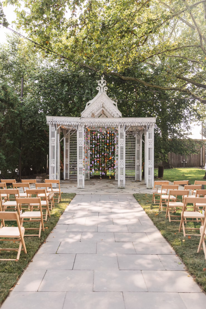Wedding ceremony arch is a large triangular structure with DIY floral curtain, surrounded by wooden chairs at Terrain in Glen Mills, PA. Taken by philly wedding photographer Madison Neumann Photography.