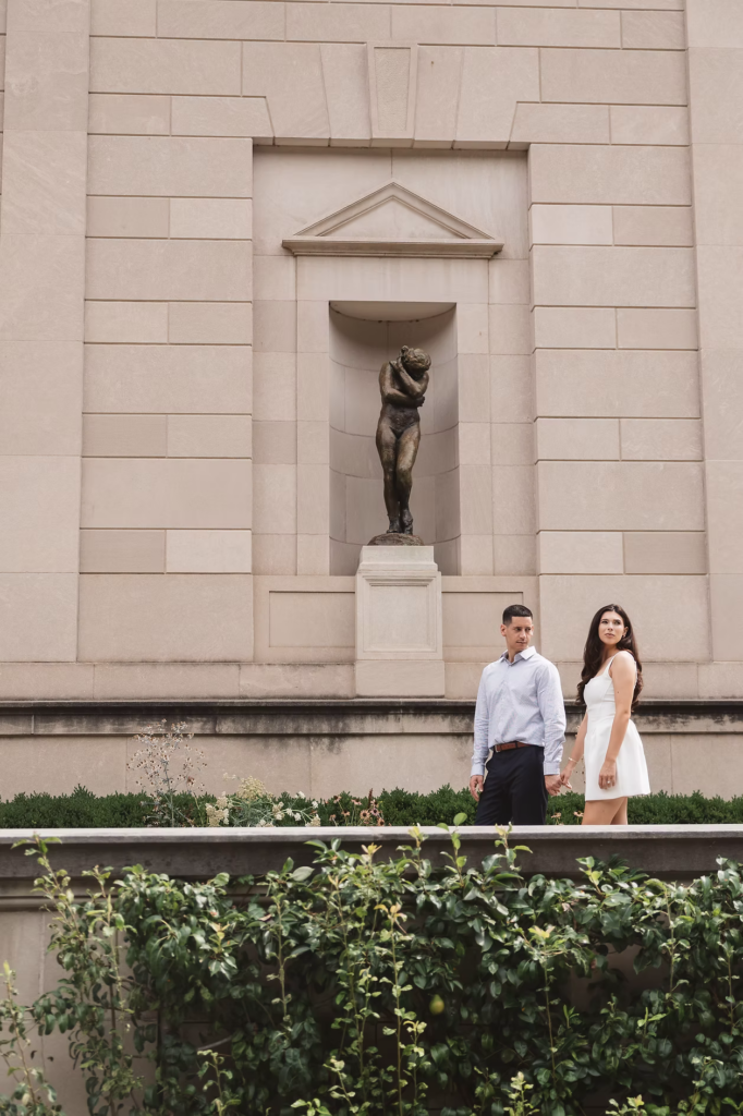 Future bride and groom stroll together by statue in garden for engagement session portraits. Photographed at The Rodin Museum in Philadelphia, PA by wedding photographer Madison Neumann Photography.