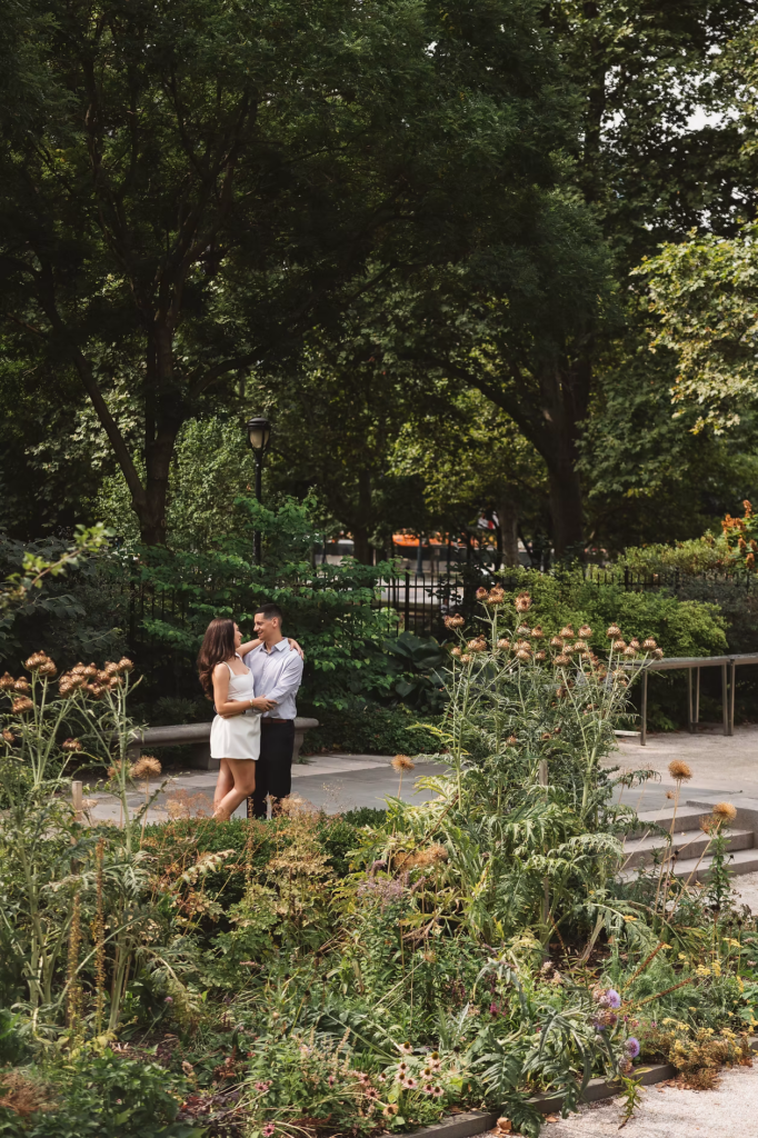 Soon to be bride and groom smile together by flowers and greenery in the garden of The Rodin Museum in Philadelphia, PA. Engagement session photographed by wedding photographer Madison Neumann Photography.