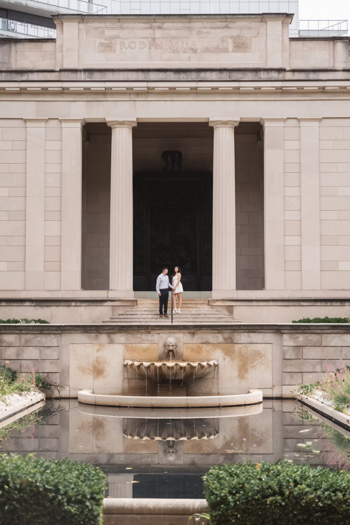 Fiancées hold hands on the steps of The Rodin Museum, sculpture water fountain and greenery surrounds them. Photographed in Philadelphia, PA by Madison Neumann Photography.