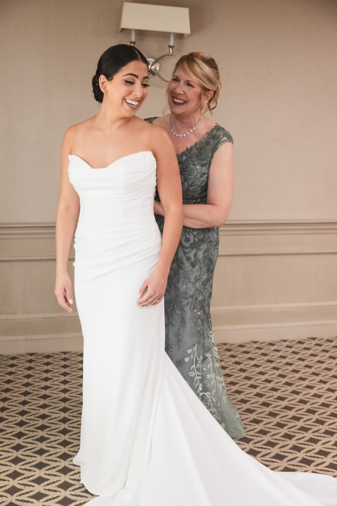 Bride smiles with her mother as she helps her get ready for her wedding day. Bride wears gown with long train, mother of the bride wears dark green detailed gown. Photographed at The Madison Hotel in Morristown, NJ by wedding photographer Madison Neumann Photography.