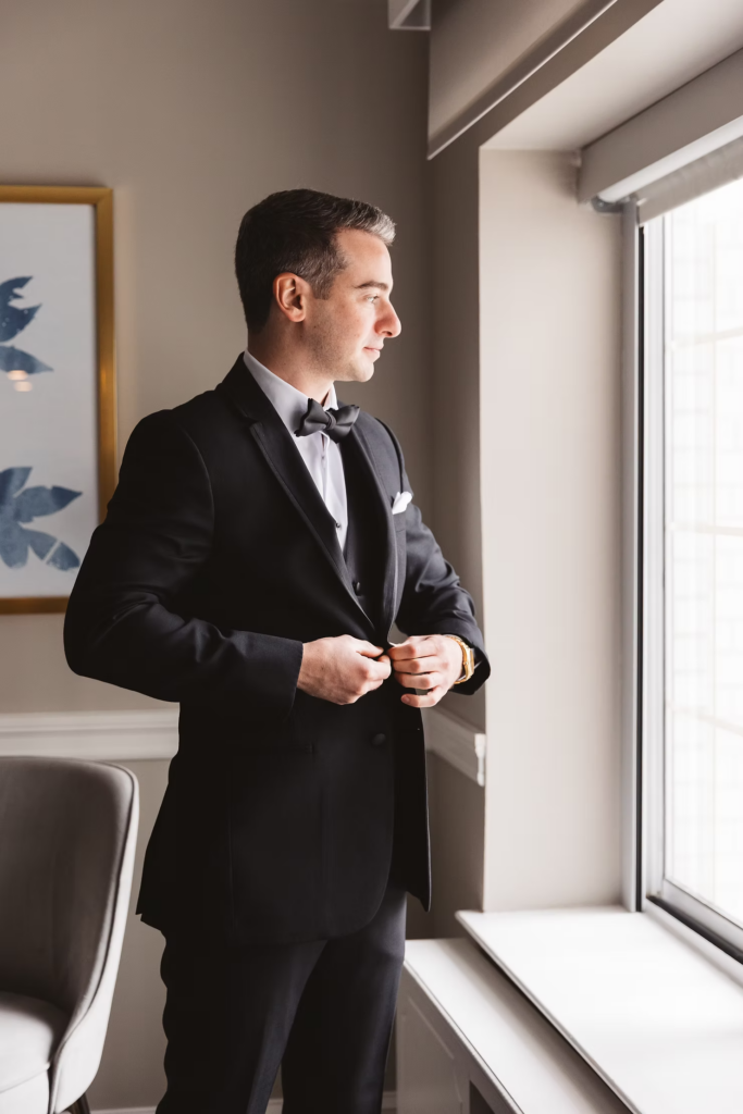 Groom, dressed in black tuxedo with bowtie, looks out the window as he gets ready for his wedding day. Photographed at The Madison Hotel in Morristown, NJ by wedding photographer Madison Neumann Photography.