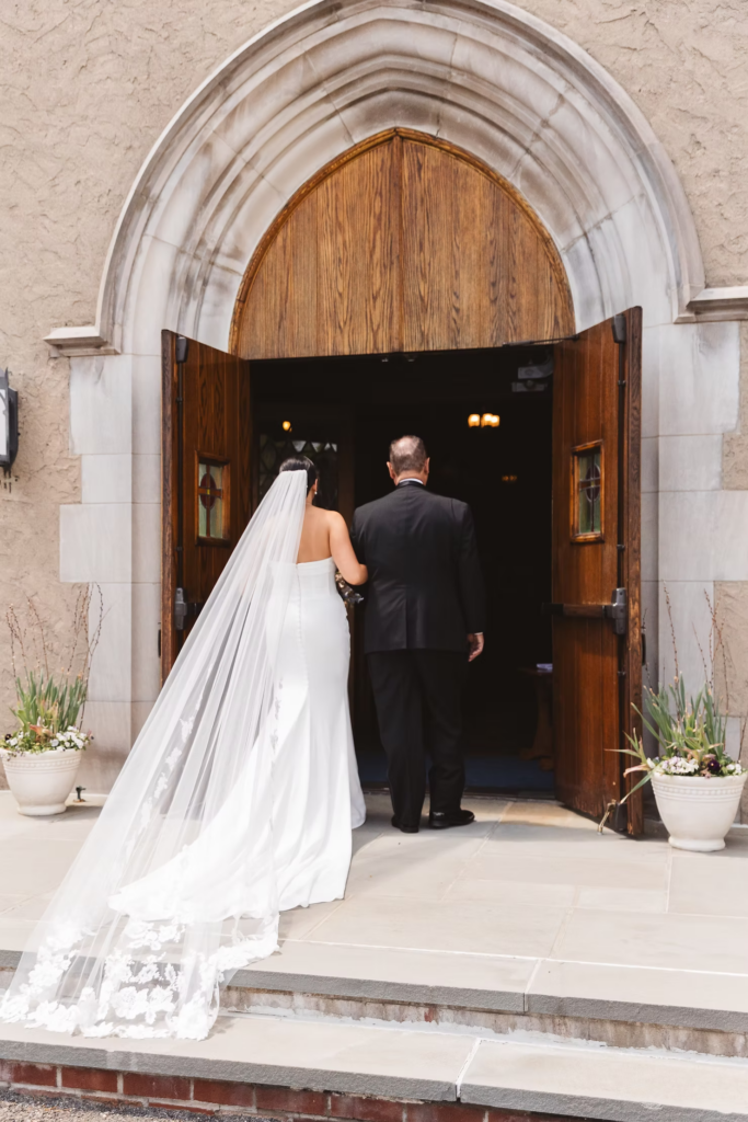 Bride holds on to her father as they enter the church for her wedding ceremony. Bride wears a long veil and gown with train. Photographed at The Madison Hotel in Morristown, NJ by wedding photographer Madison Neumann Photography.