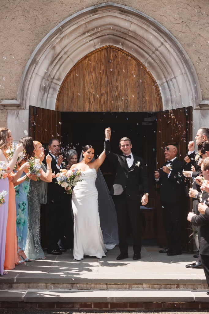 Bride and groom holding hands exiting church with guests cheering and blowing bubbles after ceremony. Photographed at The Madison Hotel in Morristown, NJ by wedding photographer Madison Neumann Photography.