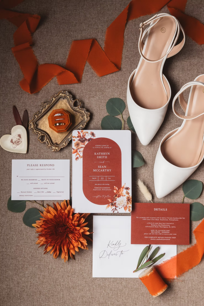 Detail photo of wedding invitation, accessories, rings, bridal shoes, ribbon, and orange flowers. Wedding day at Terrain Gardens at DelVal in Doylestown, PA, Photographed by South Jersey, Philly based wedding photographer Madison Neumann Photography.