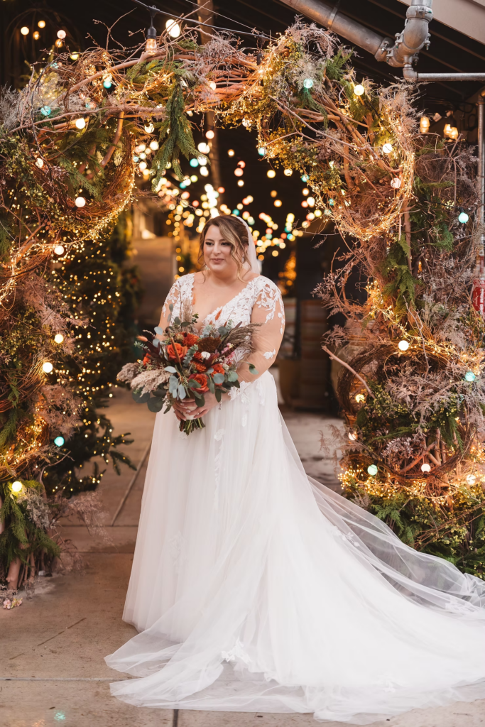 Bride in gown and veil holding bouquet under archway with string lights. Wedding day at Terrain Gardens at DelVal in Doylestown, PA, photographed by South Jersey, Philly based wedding photographer Madison Neumann Photography.