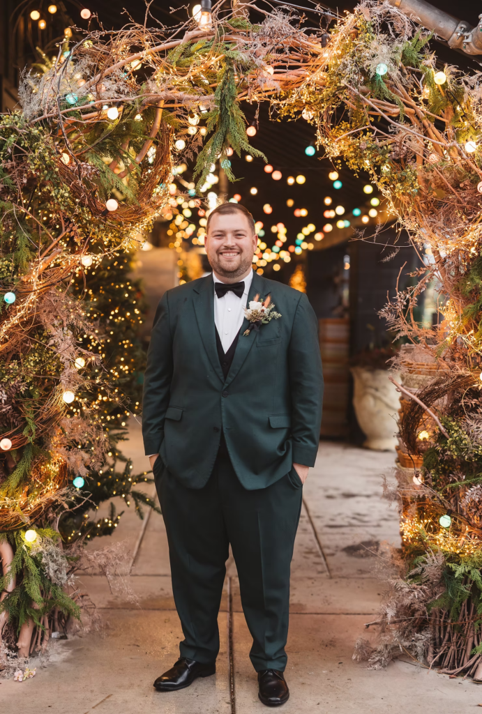 Groom in dark green tux suit and black tie under greenery archway with string lights at Terrain Gardens at DelVal in Doylestown, Pennsylvania. Wedding day photographed by South Jersey, Philadelphia based wedding photographer Madison Neumann Photography.