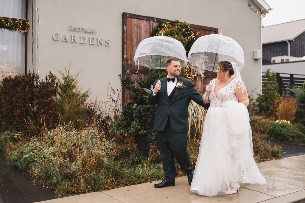 Bride and groom walk together with umbrellas under the snow on their wedding day. Photographed at Terrain Gardens at DelVal in Doylestown, PA by South Jersey, Philly based wedding photographer Madison Neumann Photography.