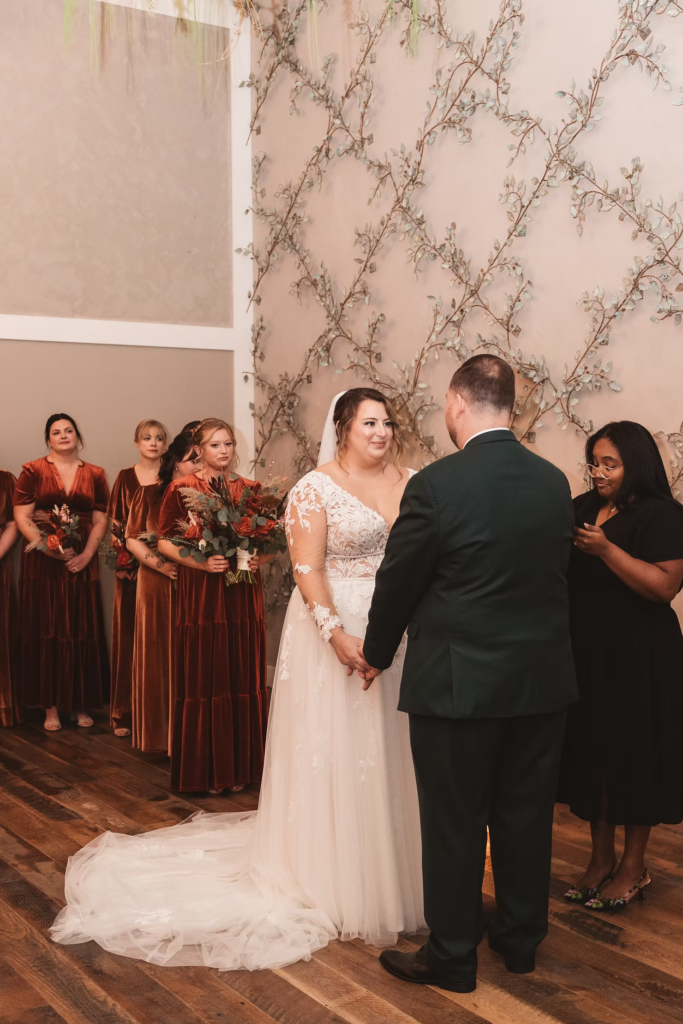Bride smiles looking at groom at the altar with bridesmaids standing behind her. Wedding day at Terrain Gardens at DelVal in Doylestown, PA, photographed by South Jersey, Philly based wedding photographer Madison Neumann Photography.