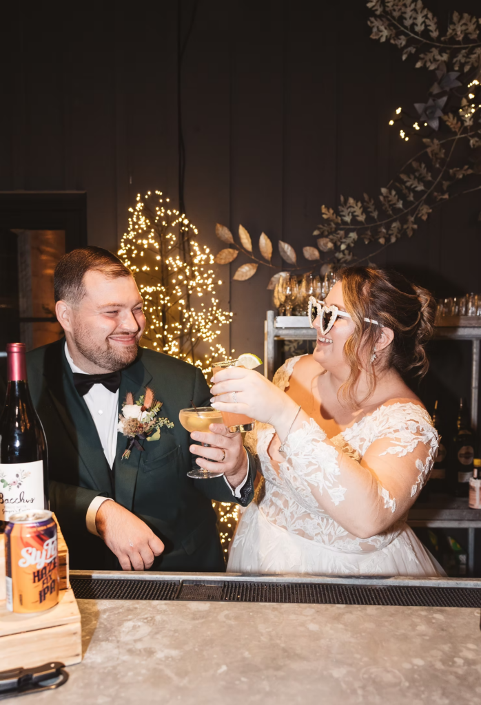 Bride wears heart-shaped sunglasses and toasts cocktail with her groom behind bar during reception. Wedding day at Terrain Gardens at DelVal in Doylestown, PA, photographed by South Jersey, Philadelphia based wedding photographer Madison Neumann Photography.