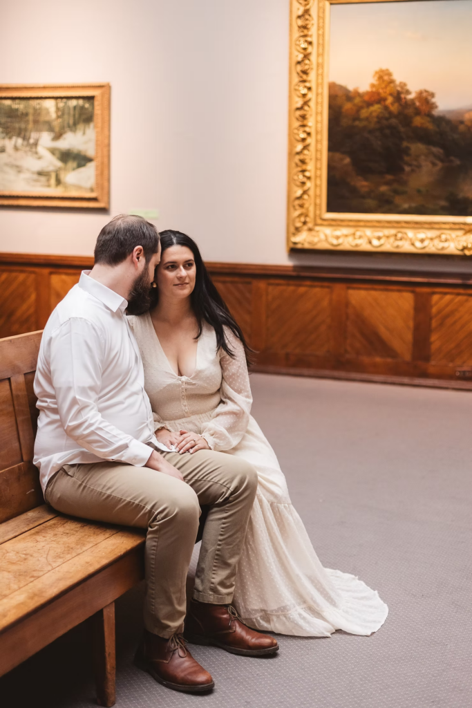 A future bride and groom sit together at the Pennsylvania Academy of The Fine Arts for their art museum engagement session in Philadelphia, looking at paintings. Photographed by Philly wedding photographer Madison Neumann Photography.