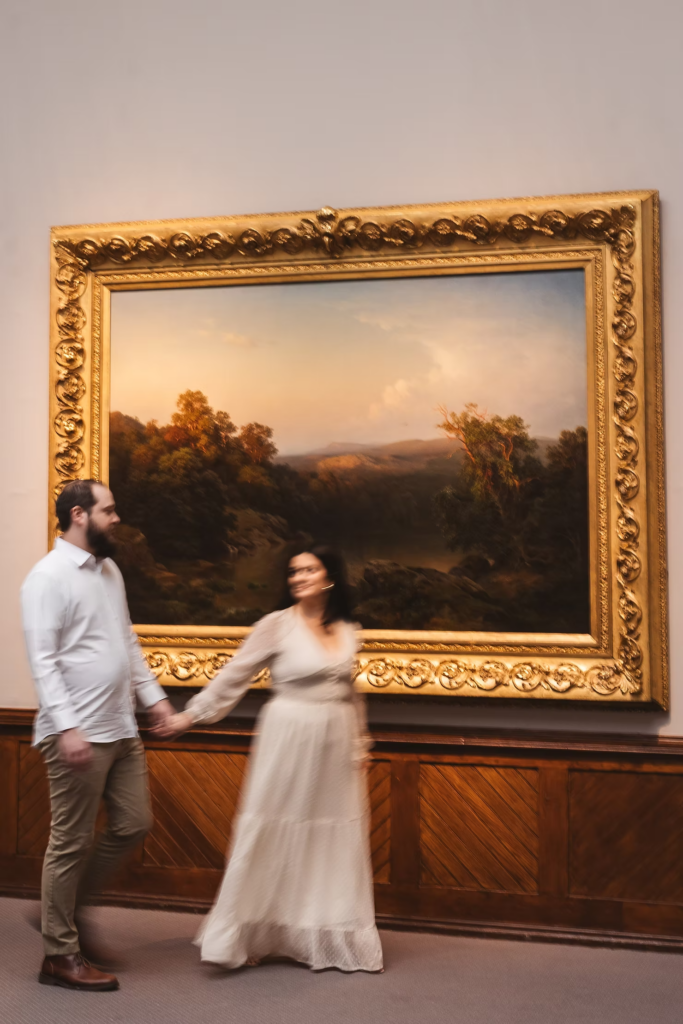 A future bride and groom walk past a large painting in the Pennsylvania Academy of The Fine Arts for their art museum engagement session in Philadelphia. Photographed by Philly wedding photographer Madison Neumann Photography.