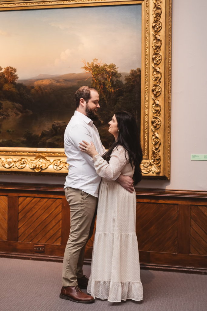 A future bride and groom pose together in front of large painting at the Pennsylvania Academy of The Fine Arts for their art museum engagement session in Philadelphia. Taken by Philly wedding photographer Madison Neumann Photography.