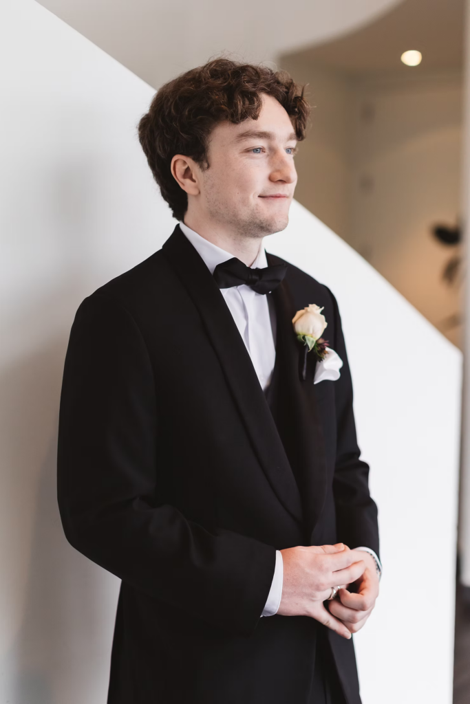 Groom in black tuxedo suit, bow tie, and boutonniere, looking out window. Wedding day at Maritime Parc in Jersey City, NJ. Photographed by South Jersey, Philadelphia based wedding photographer Madison Neumann Photography.