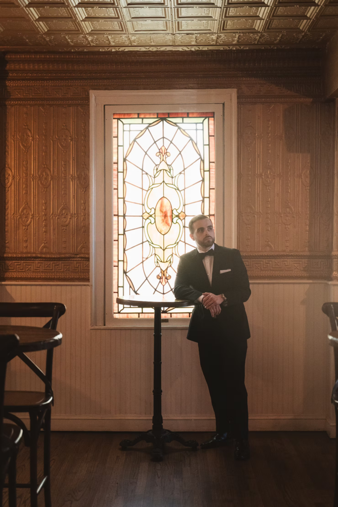 Groom in tux, black tie and suit, ready for his wedding day in front of stained glass window. Photographed at David's Country Inn in Hackettstown, NJ by South Jersey, Philadelphia based wedding photographer Madison Neumann Photography.