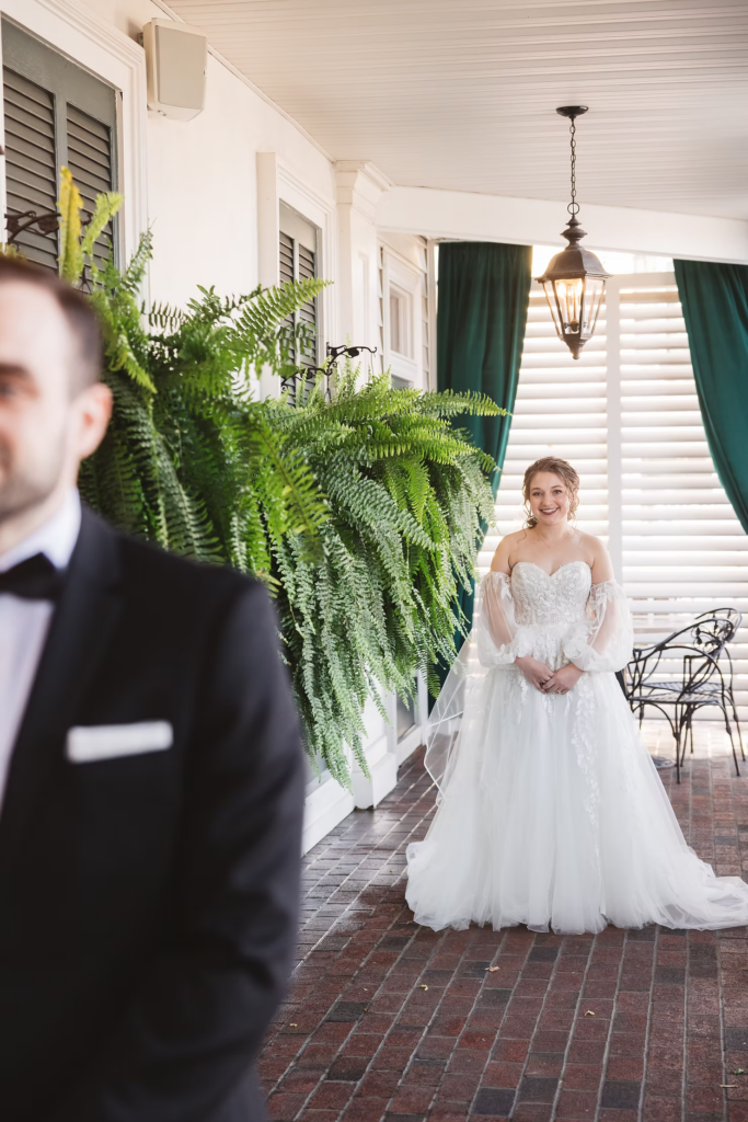 Bride waits for groom to turn around for their first look on wedding day. Standing on porch with ferns at David's Country Inn in Hackettstown, New Jersey. Photographed by South Jersey, Philadelphia based wedding photographer Madison Neumann Photography.