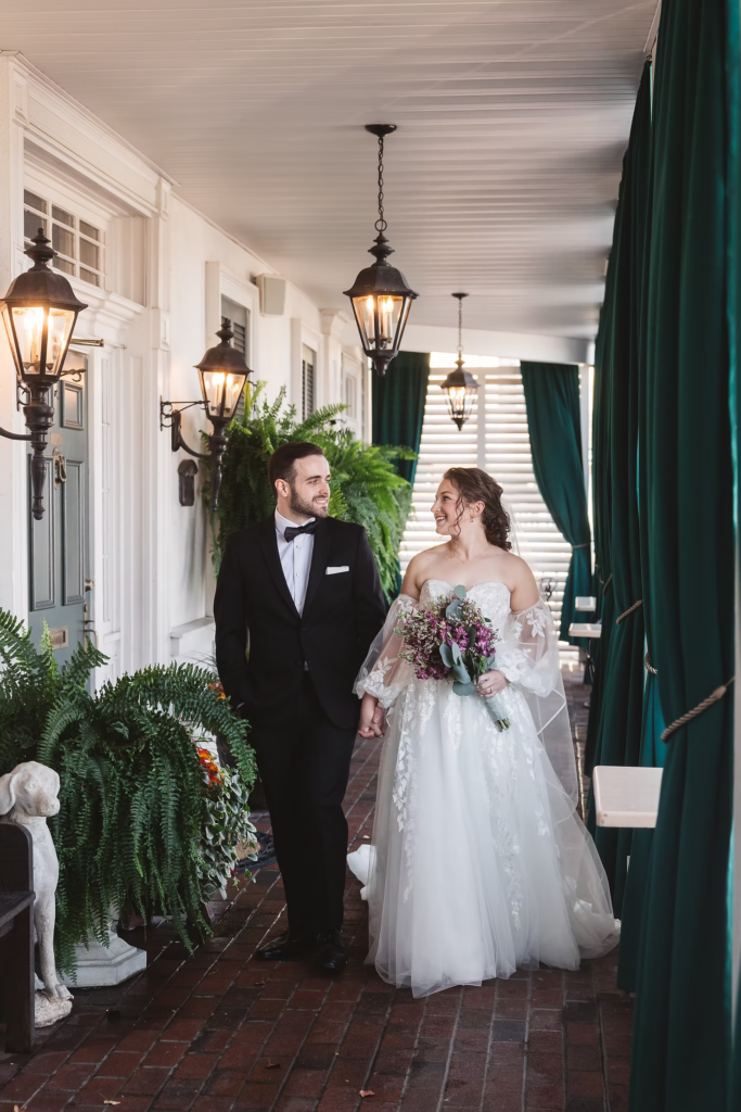 Bride and groom hold hands and walk together across porch of wedding venue, David's Country Inn in Hackettstown, New Jersey. Bride holds bouquet, porch has green curtains and plants. Photographed by South Jersey, Philadelphia based wedding photographer Madison Neumann Photography.