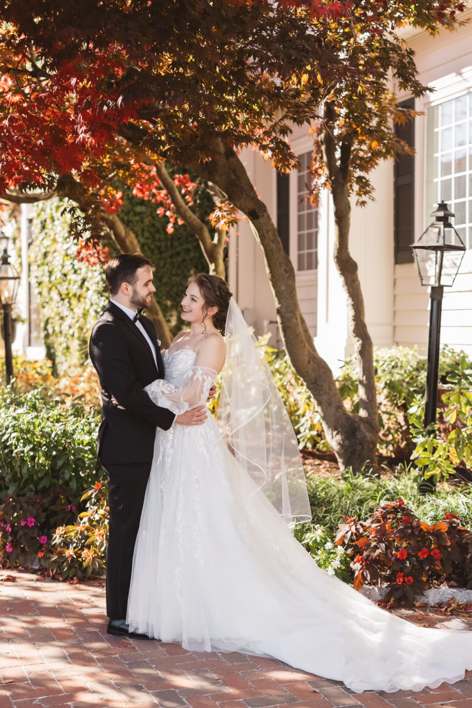Bride and groom smile together under tree with red leaves outside their wedding venue, David's Country Inn in Hackettstown, New Jersey.  Photographed by South Jersey, Philadelphia based wedding photographer Madison Neumann Photography.