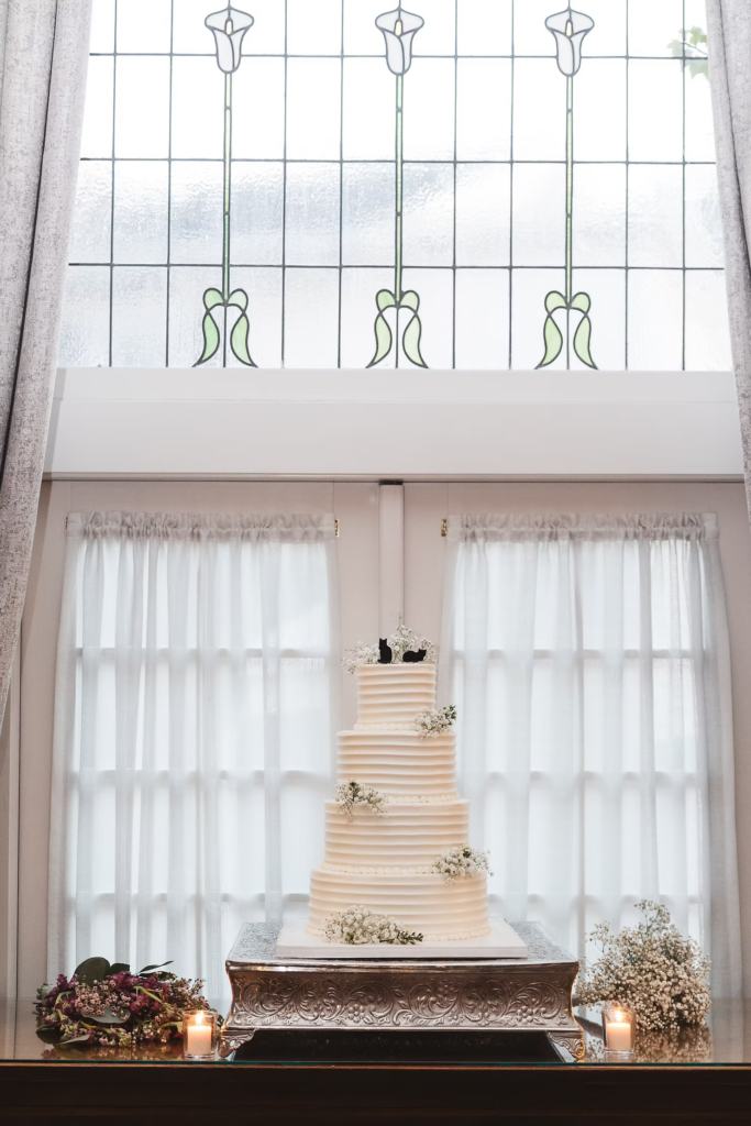 Four tier wedding cake with baby's breath and cat cake topper. Stained glass window of wedding venue David's Country Inn in Hackettstown, New Jersey. Photographed by South Jersey, Philadelphia based wedding photographer Madison Neumann Photography.