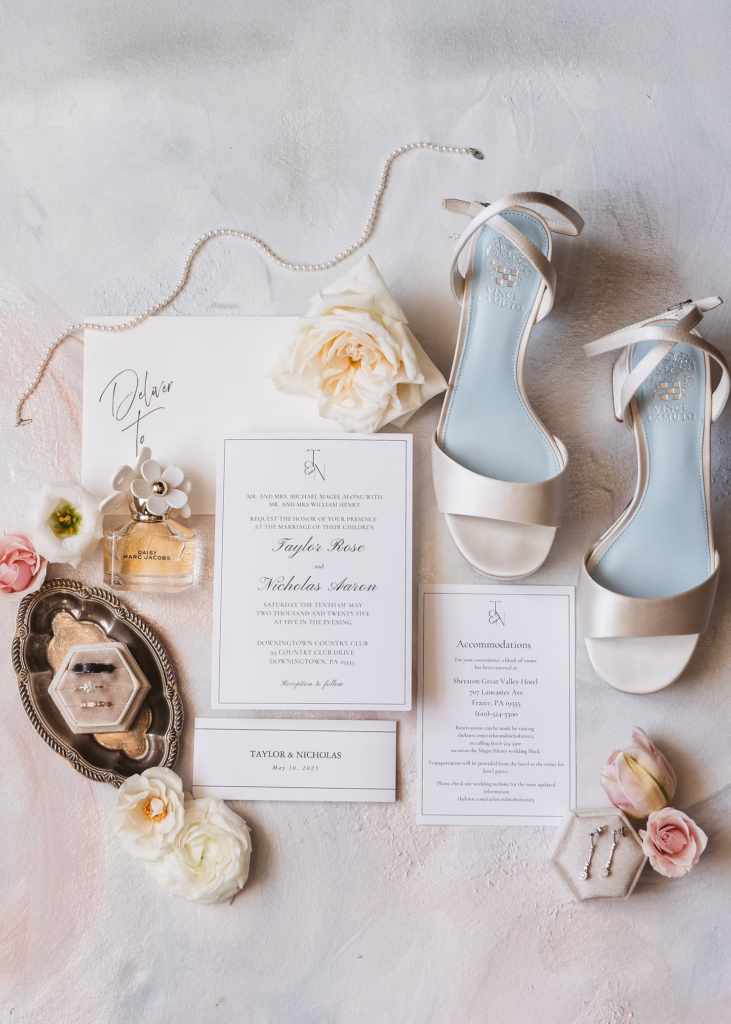 Flat lay of wedding accessories including invitation suite, pearl necklace, bridal shoes, flowers, perfume, and rings. Wedding day at Downingtown Country Club in Downingtown, PA. Photographed by South Jersey, Philadelphia based wedding photographer Madison Neumann Photography.
