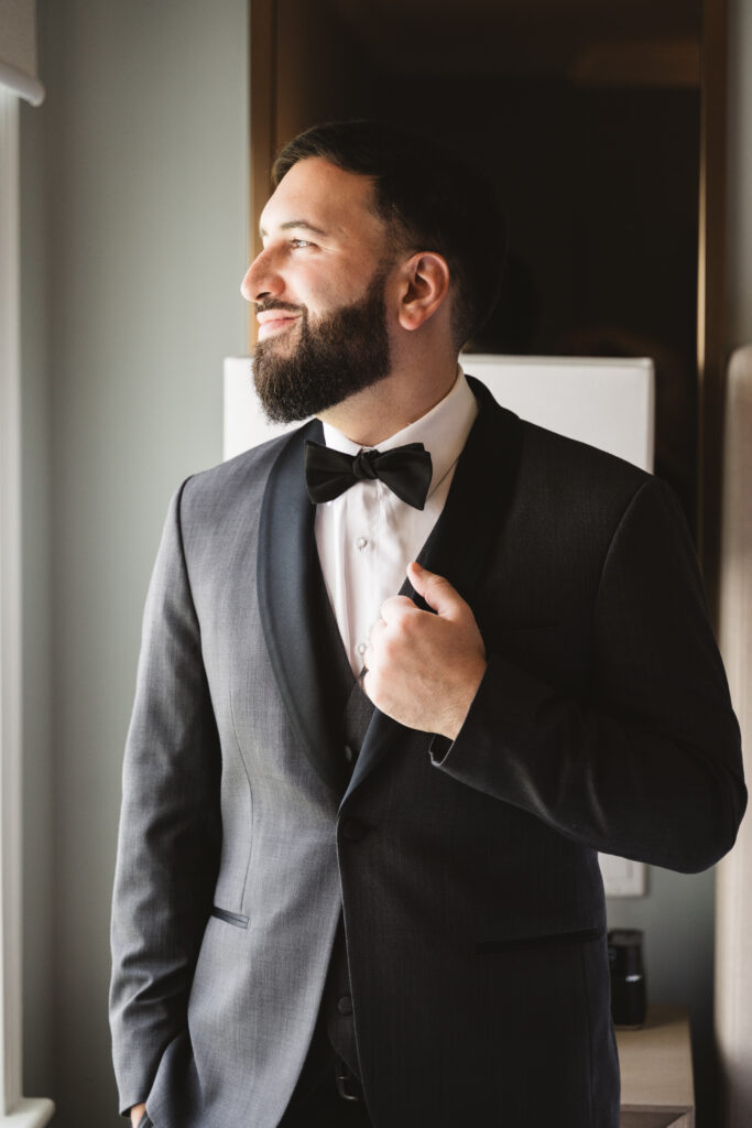 Groom smiling, wearing black tuxedo suit, looking out window. Wedding day at Blue Heron Pines Golf Club in Egg Harbor, NJ. Photographed by South Jersey/Philly based wedding photographer Madison Neumann Photography.