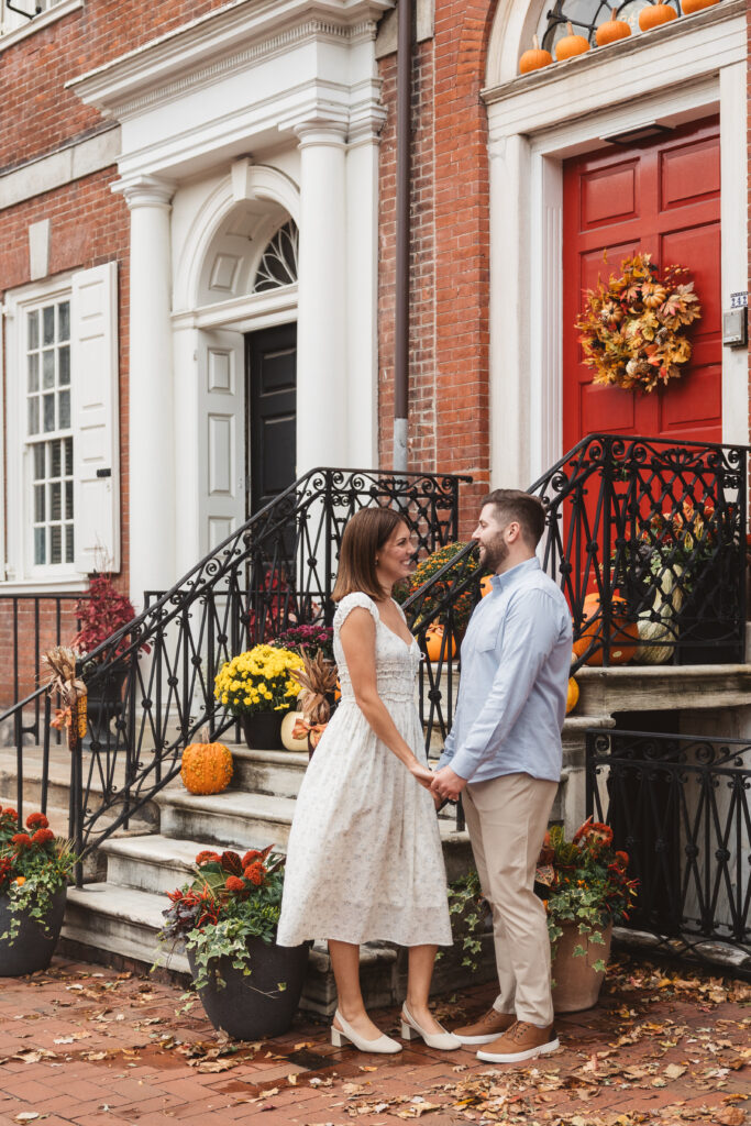 Happy couple holding hands and smiling outside homes in Philadelphia that are decorated for fall with wreaths and pumpkins, photographed by Philadelphia and New Jersey based travel wedding photographer, Madison Neumann Photography.