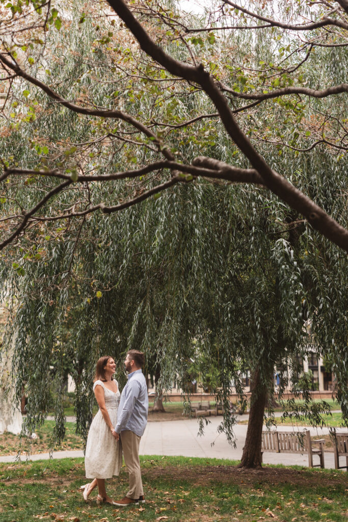 Wide shot of couple holding hands under a tree in the park, smiling at one another, photographed by Philadelphia and New Jersey based travel wedding photographer, Madison Neumann Photography.