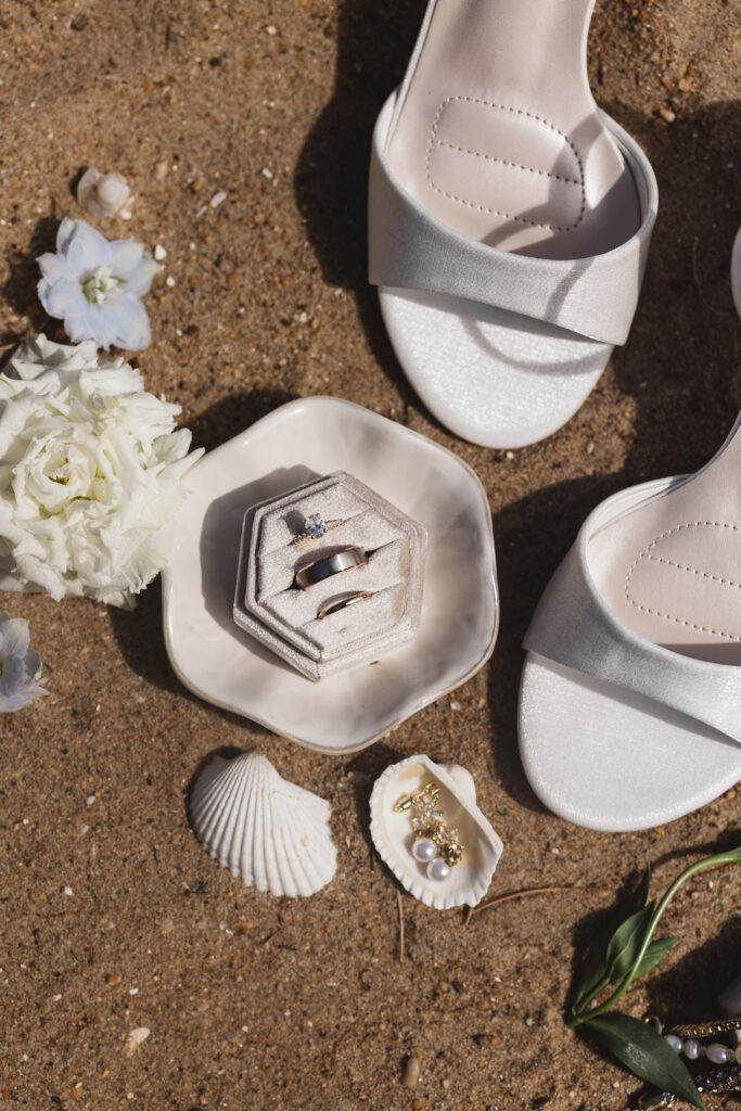 Flat lay of rings, flowers, and heels on sand in Outer Banks, North Carolina photographed by New Jersey and Philadelphia based travel wedding photographer, Madison Neumann Photography.