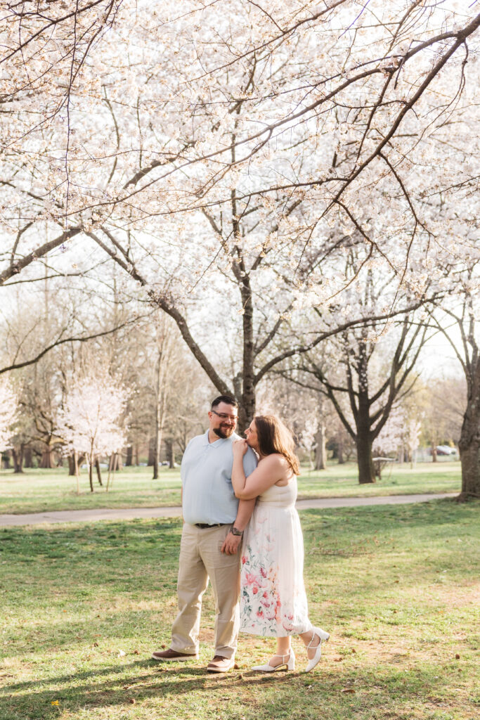 Bride stands behind groom and leans her head on his shoulder as they stand in the middle of the park with the blossom trees matching the florals on the bottom of the brides dress, photographed by Philadelphia and New Jersey based travel wedding photographer, Madison Neumann Photography.