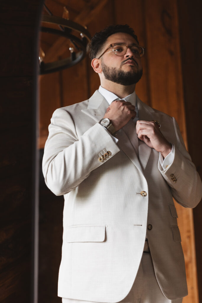 Groom in a cream suit is looking in the mirror and fixing his tie, photographed by Philadelphia and New Jersey based travel wedding photographer, Madison Neumann Photography.