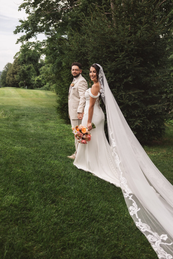 Groom holding the brides hand and they both look back at the camera on the grass with the bride's long lacy veil. Photographed by Philadelphia and New Jersey based travel wedding photographer, Madison Neumann Photography.