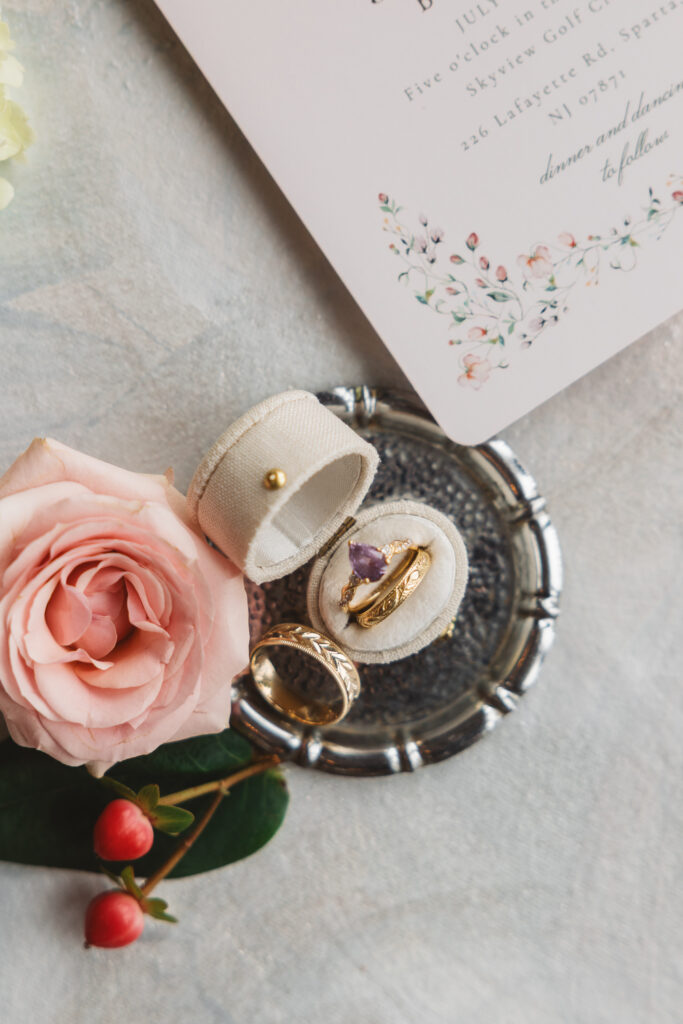 Bride and Grooms rings on a silver tin with a pink rose at Skyview Golf Club in Sparta New Jersey, photographed by Philadelphia and New Jersey based travel wedding photographer, Madison Neumann Photography.