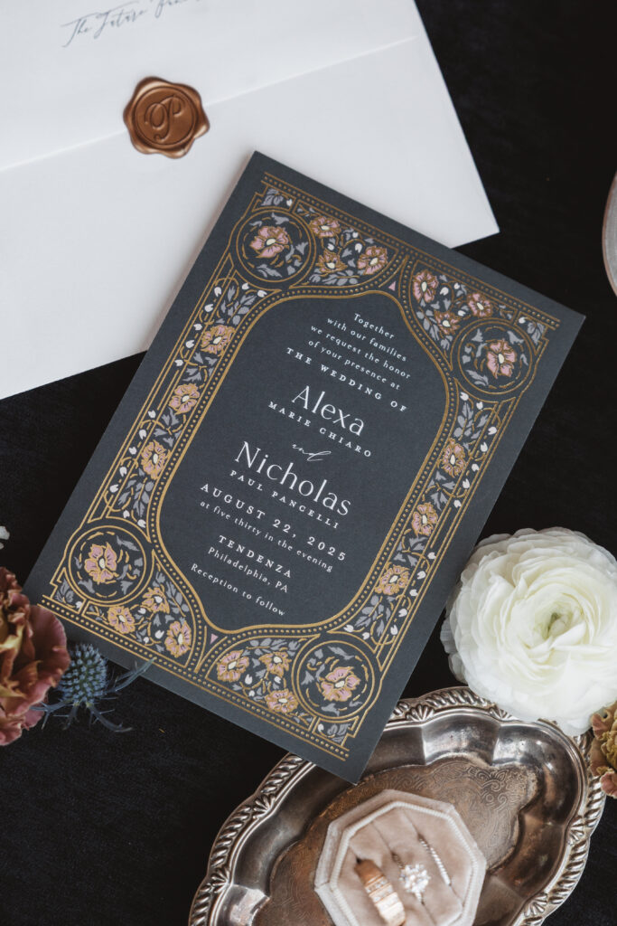 Flat lay of wedding invitation on a dark background with flowers and the rings photographed by Philadelphia and New Jersey based travel wedding photographer, Madison Neumann Photography.