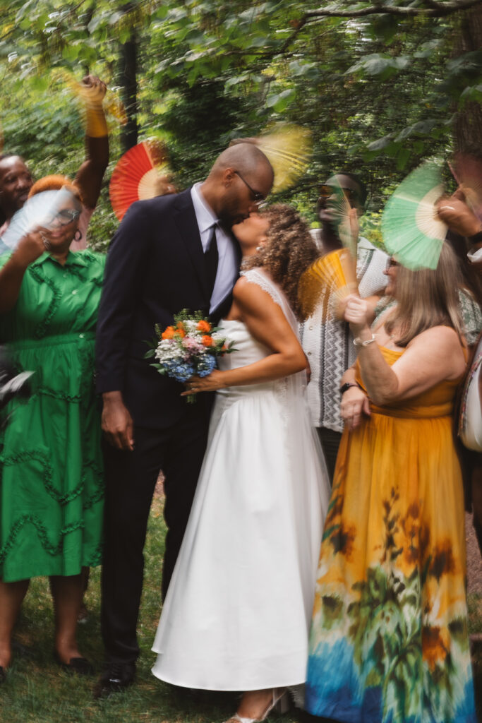 Blurred zoomed in photo of the couple kissing while surrounded by loved ones and blurred fans waving, photographed by Philadelphia and New Jersey based travel wedding photographer, Madison Neumann Photography.