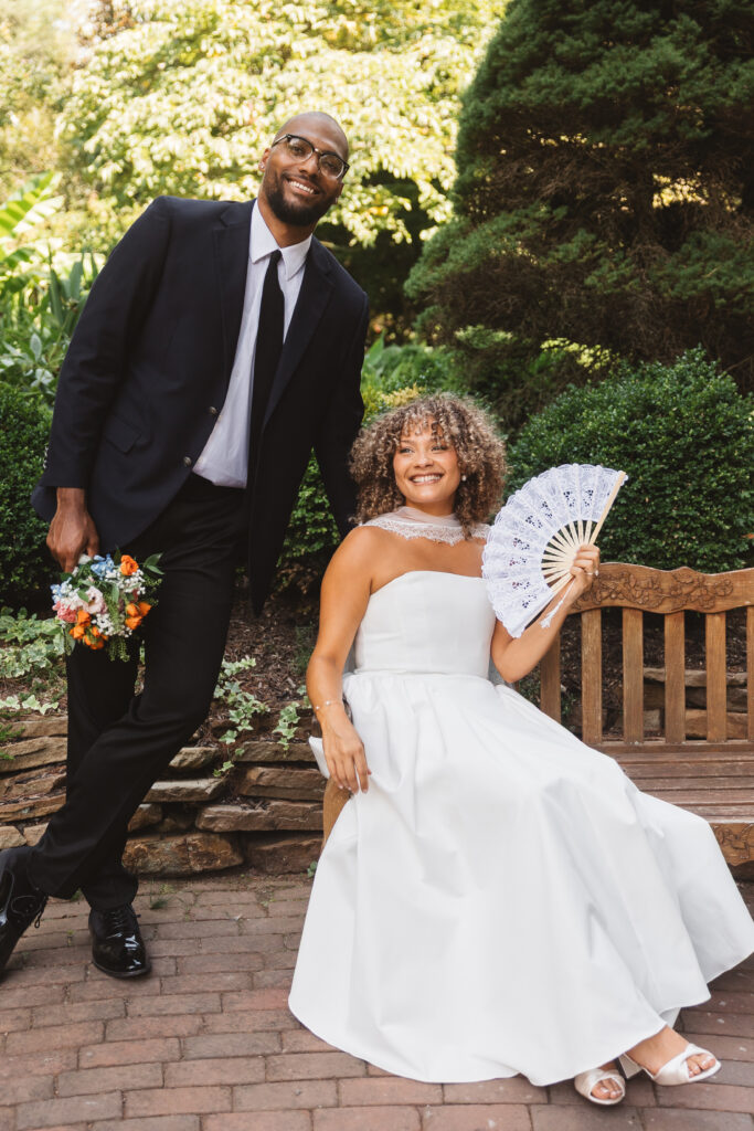 Bride is pictured on a bench with her fan as the groom leans against the bench while standing and holding her bouquet, photographed by Philadelphia and New Jersey based travel wedding photographer, Madison Neumann Photography.